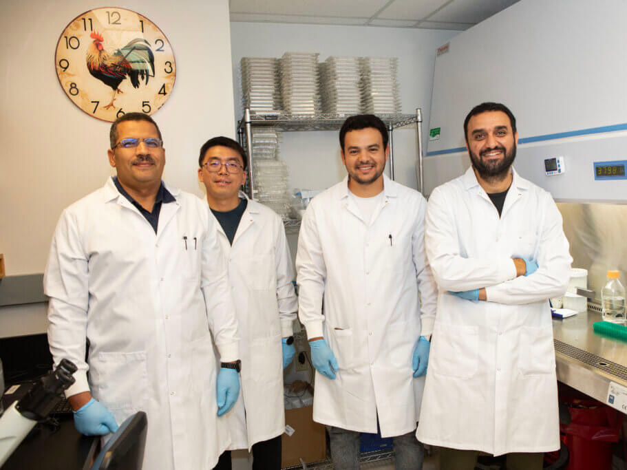 Four scientists in white lab coats and blue gloves pose in a laboratory beside equipment and shelves of containers.